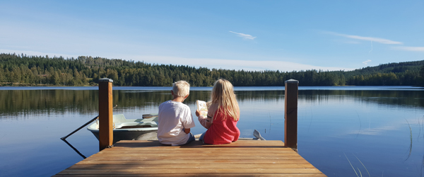 Två barn läser en bok på bryggan vid en sjö en solig sommardag. Foto: Hampus Karlsson
